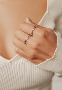 Bague en argent au design tordu avec de petits accents de feuilles. Portée sur une main ayant un teint de peau clair, sur un pull crème texturé.