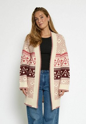 Strickjacke in Creme mit geometrischen Mustern in Pink und Burgunder, offene Vorderseite, weite Ärmel und gerippte Bündchen, kombiniert mit blauen weiten Jeans.