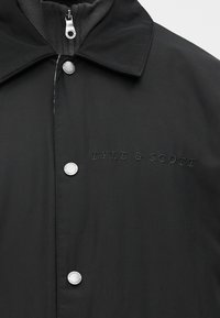 Casaco preto resistente à água com gola clássica, apresenta fecho de botão de pressão prateado e logótipo bordado "Lyle & Scott" no peito.