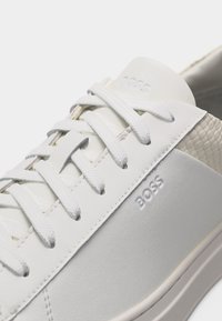 Κοντινή λήψη λευκού αθλητικού παπουτσιού BOSS που δείχνει την υφή στη φτέρνα, το μπροστινό μέρος με κορδόνια και το ανάγλυφο όνομα της μάρκας στο πλάι και στη γλώσσα.