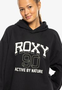 Schwarzer Hoodie aus einem weichen Material mit einem großen weißen Grafikdesign, auf dem "ROXY 90" und darunter "ACTIVE BY NATURE" in kontrastierenden Farben aufgedruckt sind.