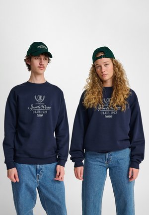 Marineblauwe sweatshirt met geribbelde manchetten en tailleband, met witte tekst en logo. Model draagt lichte spijkerbroek en een groene pet.