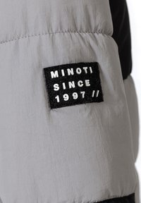 Pelēka puffa jaka ar melnu plāksnīti, uz kuras ir balti izcirsti burti "MINOTI SINCE 1997 //". Audums ir gluds ar nelielu spīdumu.