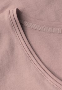 Světle růžová bavlněná látka s ribovaným kulatým výstřihem. Má ploché švy a jemnou texturu, což podtrhuje její jednoduchý, klasický design.