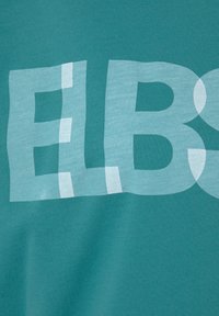 Türkises Baumwoll-T-Shirt mit großen, halbtransparenten weißen Buchstaben, die "ELBS" lesen, sowie unregelmäßigen Formen und weicher Textur.