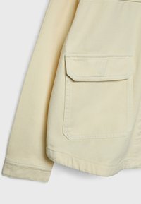 Détail d'une veste crème à manches longues montrant une grande poche avant avec rabat et coutures visibles.