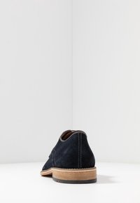 Chaussure en suède bleu marine avec un bout arrondi, des détails cousus, une doublure en cuir marron et une semelle en caoutchouc beige clair. Design moderne et minimaliste.