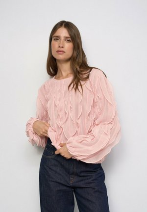 Mujer con cabello largo y castaño que lleva una blusa rosa claro texturizada con volantes y vaqueros azul oscuro de talle alto sobre un fondo blanco liso.