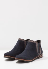 Botas de ante azul marino hasta el tobillo con ribete texturizado marrón y tacones de madera bajos, ubicadas sobre una superficie blanca con un fondo sencillo.