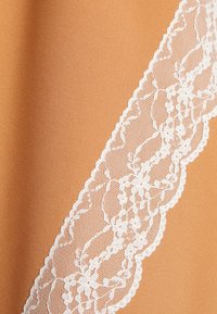 Bordure en dentelle florale blanche avec des bords festonnés sur un fond de tissu lisse couleur sable, montrant des textures et des motifs détaillés.