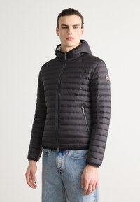 Čierna nadýchavaná bunda s kapucňou, prešívaný dizajn, predný zips, bočné vrecká a logo na ľavom rukáve. Nosené s svetlomodrými džínami.