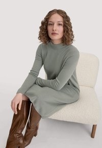 Hellgrünes geripptes Kleid mit hohem Kragen und langen Ärmeln, kombiniert mit kniehohen schokoladenbraunen Lederstiefeln, sitzt auf einem strukturierten weißen Stuhl.