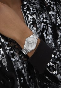 Silberne Uhr mit einem runden Ziffernblatt, kompliziertem Innendesign und Strassakzenten an einem Metallarmband, getragen an einer Hand gegen eine Paillettenjacket.