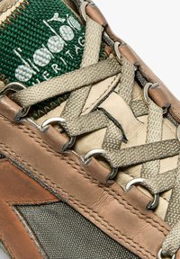 Zapatillas en tono marrón y verde con parte superior de tela texturizada, cordones beige, ojales metálicos y un logo en la lengüeta. Presenta elementos de diseño en capas.