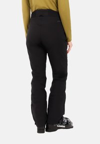 Zwarte skibroeken met een hoge, elastische tailleband. De stof is glad en er zitten ritsen aan de onderbenen. Gemaakt om te dragen met zwarte skischoenen.