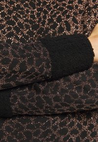 Pull noir avec un motif léopard en or rose brillant. Il présente des poignets en polaire noire texturée et des détails complexes sur tout le tissu.