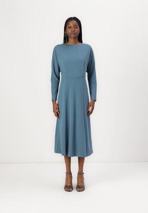 Teal long-sleeve dress with a boat neckline, fitted bodice, and flared skirt. Accessorized with black strappy heels. Smooth fabric texture.