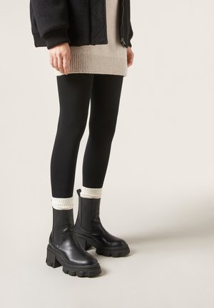 Svarta ankelskängor med kraftiga, räfflade sulor och elastiska sidopaneler, tillsammans med svarta leggings och krämliknande stickade manschetter.