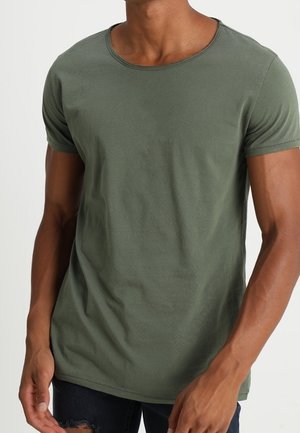 Hombre con una camiseta ajustada de manga corta y cuello redondo de color verde oliva y vaqueros oscuros rotos, de pie frente a un fondo liso.