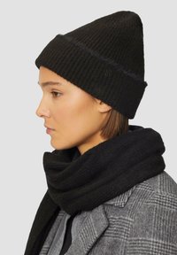 Zwarte geribbelde gebreide beanie met een puntige top en subtiele glans, gecombineerd met een zachte zwarte sjaal, gedragen over een grijs geruit jasje.