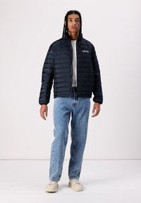 Sort pufferjakke med vandret quiltet design, høj krave og lynlås foran. Bæres med lyseblå jeans og cremede sneakers.