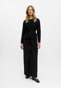 Haut peplum noir avec des manches longues et une taille ajustée, associé à un pantalon noir à jambes larges et des chaussures sombres, sur un fond neutre.