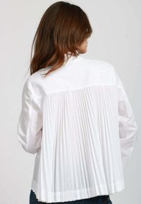Blusa bianca con schiena plissettata, maniche lunghe e colletto. Il tessuto ha una texture liscia, con un design classico e sartoriale.