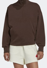 Sudadera de cremallera marrón hecha de una tela suave, con mangas largas y un corte relajado. Combinada con pantalones cortos crema y un detalle de logo rojo.