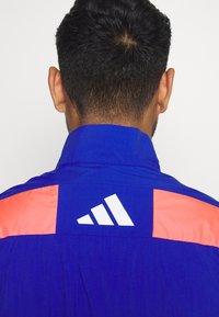 Chaqueta deportiva azul y naranja con cuello alto. Presenta un logo blanco en la parte trasera y tela suave y ligera.