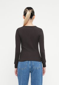 Vrouw met een blonde paardenstaart die een aansluitend zwart shirt met lange mouwen en hoge taille blauwe jeans draagt, met de rug naar de camera tegen een witte achtergrond.