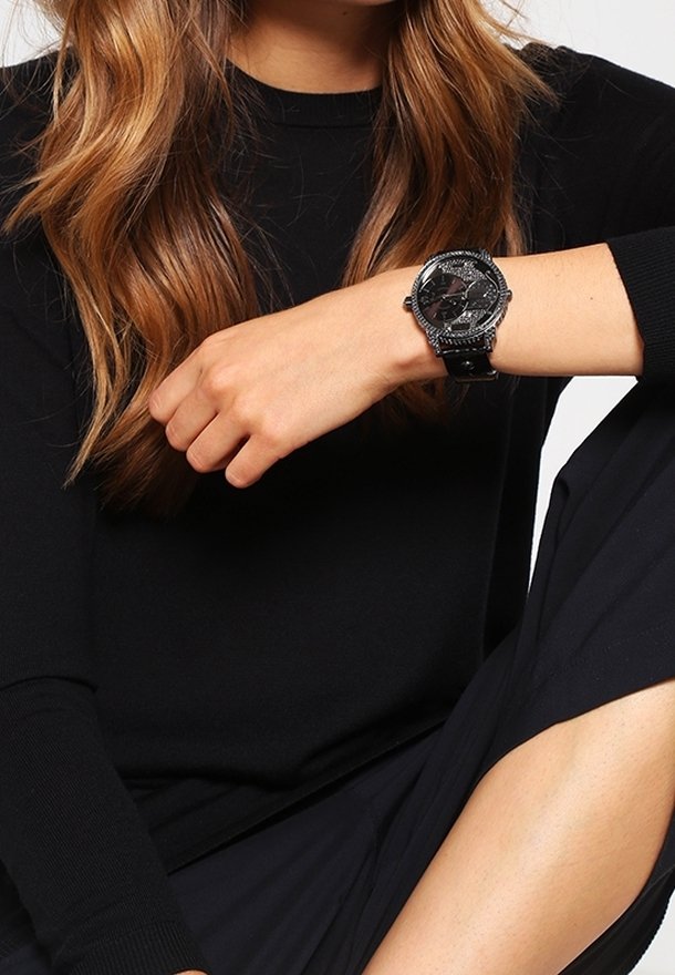 Femme aux cheveux châtain clair portant un haut à manches longues noir et un pantalon, montrant une montre noire au poignet gauche.