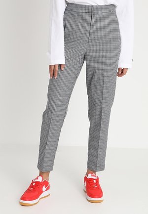 Unterkörper einer Person, die schwarz-weiß karierte Hose, weißes Langarmshirt und rot-weiße Nike-Sneaker vor weißem Hintergrund trägt.