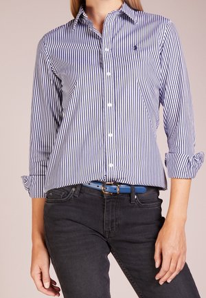 Femme portant une chemise rayée verticale bleue et blanche à boutons, un jean noir et une ceinture bleue avec une boucle dorée, debout, les bras détendus.