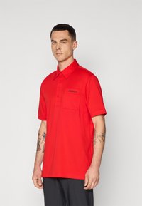Tricou polo roșu realizat dintr-un material neted, cu guler, placket cu butoane și un buzunar pe piept cu logo. Mâneci scurte, croială lejeră.