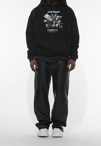 Svart hoodie med grafisk design som visar kontanter och en kompass. Kombinerad med svarta läderbyxor och vita sneakers, stående på en enkel bakgrund.