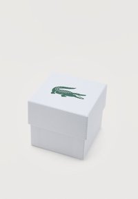 Λευκός υφασμάτινος κουτί με πράσινο λογότυπο κροκοδείλου στο καπάκι, σε απλό λευκό φόντο.