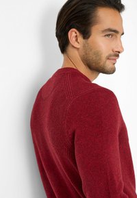 Maglione rosso lavorato a maglia con una texture a coste, scollatura rotonda e dettagli distintivi sulle cuciture delle spalle. Indossato da una persona di profilo.