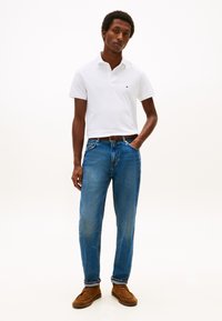 Hvit kortermet poloshirt med en liten logo, kombinert med blå jeans med rette ben og brune slip-on sko. Enkel design og passform.