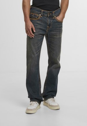 Mężczyzna w wyblakłych, ciemnoniebieskich prostych dżinsach i białych trampkach, stojący z jedną ręką w kieszeni na jednolitym tle.