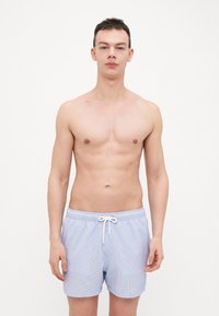 Modré a biele pásikavé plavkové šortky s elastickým pásom a detailom šnúrky, vystavené na modeli bez vrchného oblečenia.