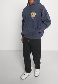 Sudadera con capucha azul marino con bolsillo frontal, diseño gráfico que presenta un sol y una cara. Combinada con pantalones negros y zapatillas blancas.