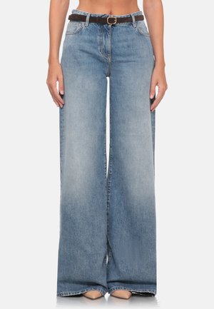 Donna che indossa jeans azzurri a gamba larga con cintura marrone scuro e scarpe beige appuntite su fondo bianco.