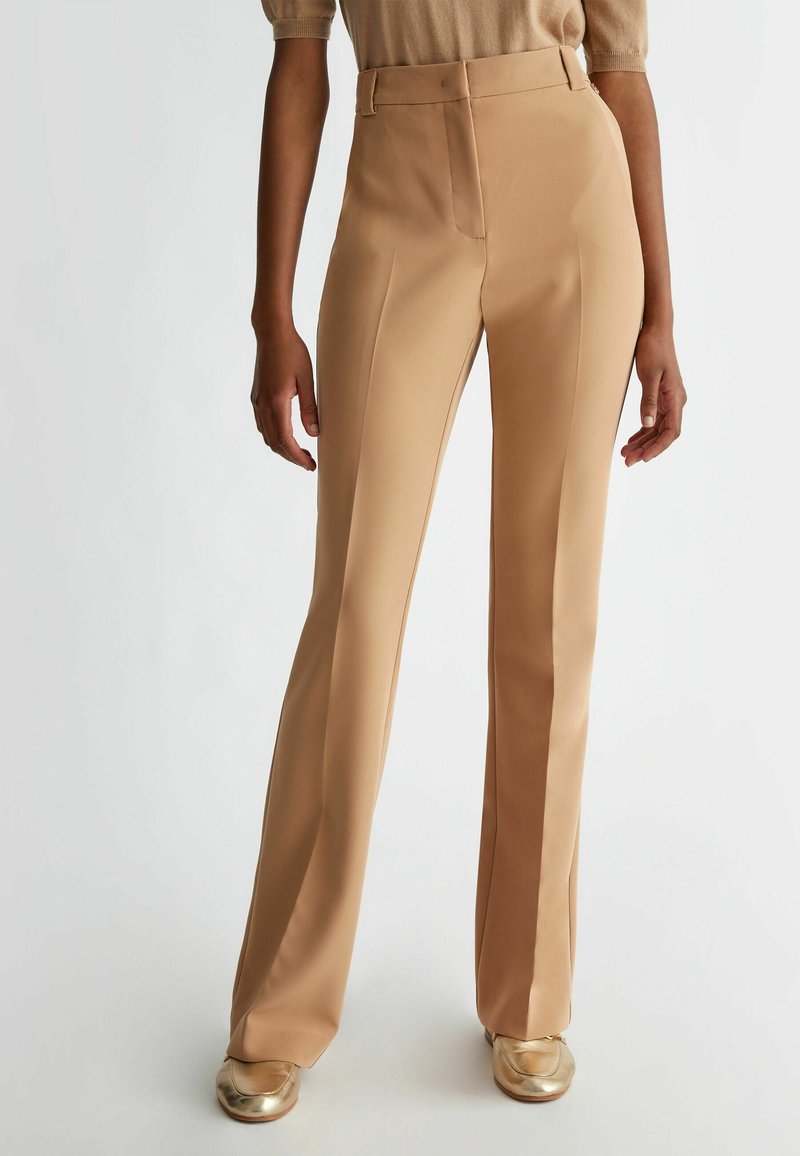 Beige op maat gemaakte broeken met een hoge taille, gestructureerde stof, recht pijp ontwerp en minimale details. Gecombineerd met gouden schoenen.