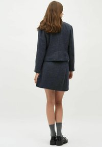 Marineblauwe tweedanzug met een getailleerde blazer en een bijpassende rok. De blazer heeft een cropped snit; de rok is knielengte. Geaccentueerd met grijze sokken en zwarte schoenen.