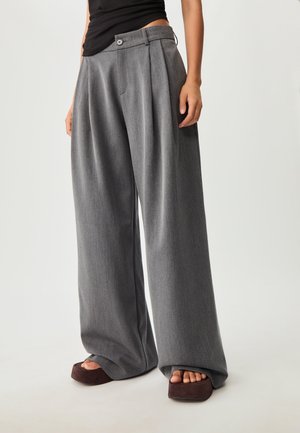 Weit geschnittene Hosen aus einem strukturierten grauen Stoff mit hoher Taille, Falten und einem Knopfverschluss. Kombiniert mit klobigen Plateausandalen.
