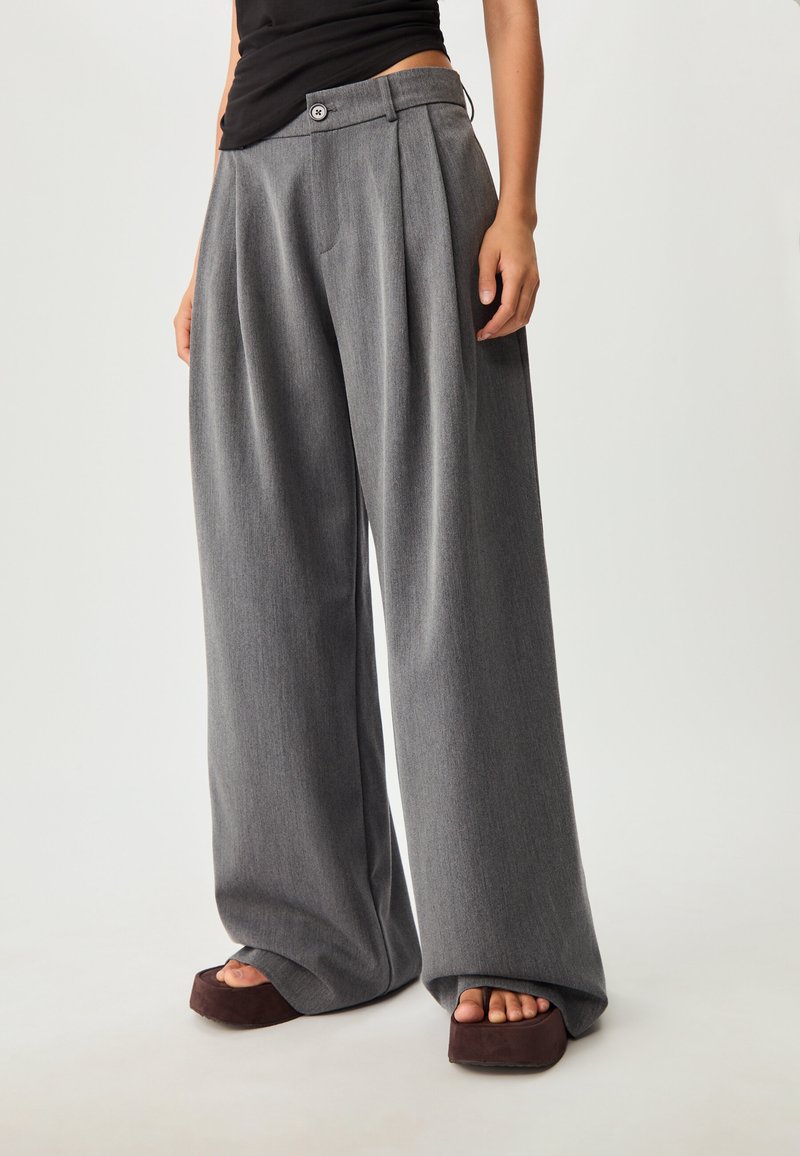 Weit geschnittene Hosen aus einem strukturierten grauen Stoff mit hoher Taille, Falten und einem Knopfverschluss. Kombiniert mit klobigen Plateausandalen.