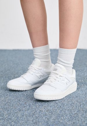 Nogi w białych prążkowanych skarpetach do kostki i białych sneakersach New Balance stojące na niebieskim dywanie.