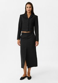 Schwarze Cropped-Blazer mit langen Ärmeln, fünf Knöpfen und zwei vorderen Taschen. Passender schwarzer Midi-Rock mit einem Schlitz auf der Vorderseite. Schwarze flache Schuhe.