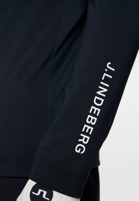 Maglia a manica lunga blu marina in tessuto liscio, con il testo bianco "J. Lindeberg" lungo la manica. Include un guanto con accenti bianchi e neri.