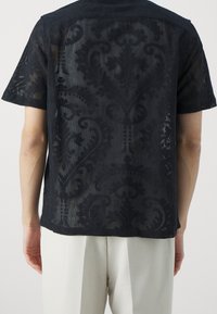 Camicia nera trasparente a maniche corte con un motivo damascato testurizzato e taglio dritto, abbinata a pantaloni di colore chiaro.